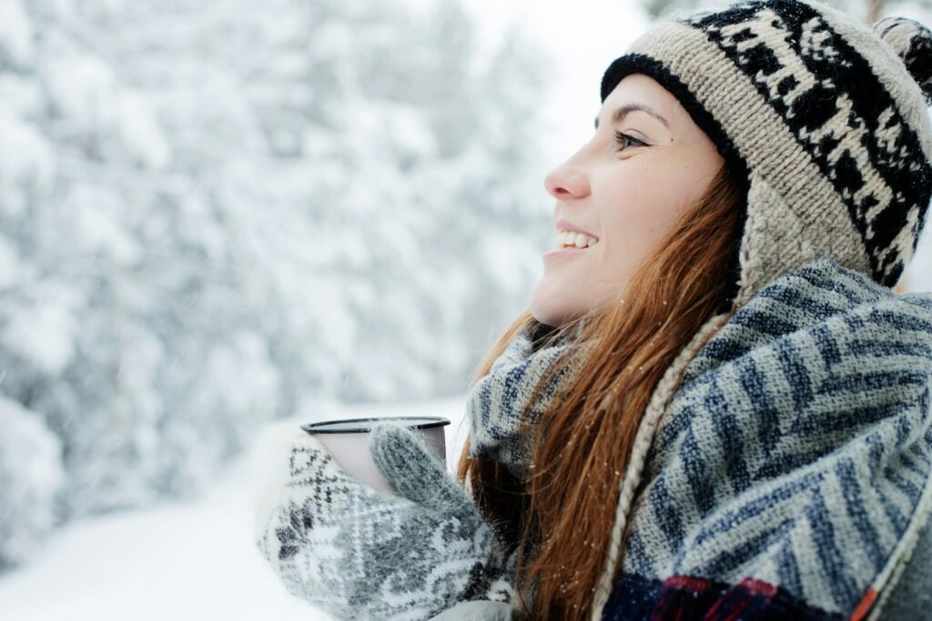 A woman in winter attire smiles while holding a mug in a snowy outdoor setting.