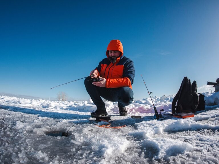 Angler crouching beside an ice hole while setting up fishing gear during ice fishing in Banff, surrounded by snow-covered ice and clear blue winter sky.