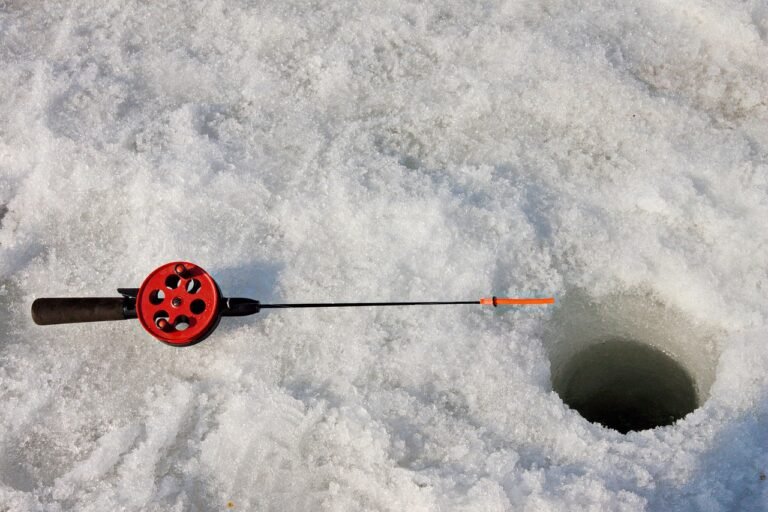Close-up of an ice fishing hole drilled into a frozen lake during ice fishing in Banff, with boots, fishing rod, and clear ice visible.
