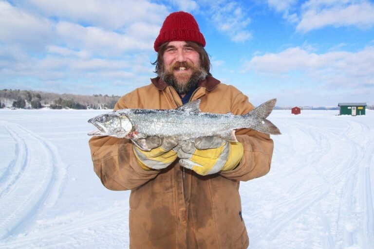 Smiling angler holding a freshly caught fish on a frozen lake during ice fishing in Banff, with snow-covered ice huts and winter landscape in the background.