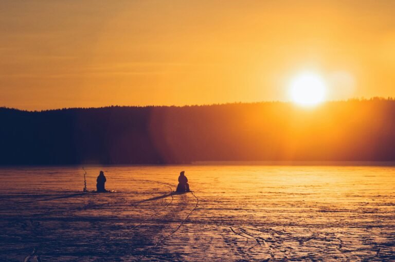 Silhouetted figures ice fishing on a frozen lake during a vibrant sunset in Lahti, Finland.