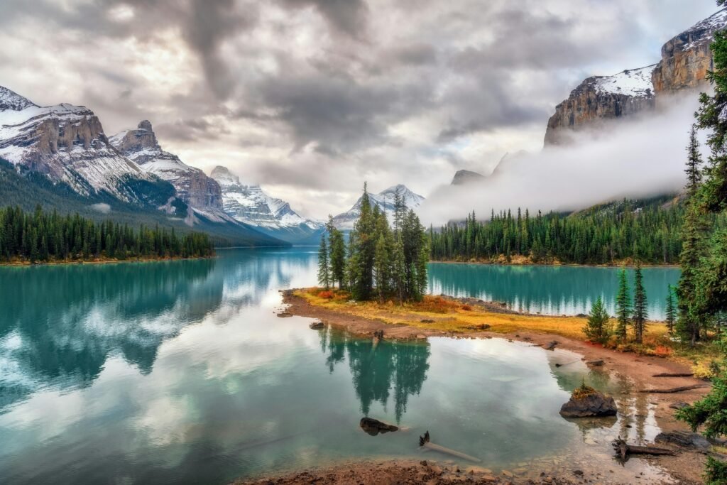 Turquoise mountain lake with pine trees on a small peninsula, misty clouds, and rugged snow-dusted peaks, capturing the serene wilderness often compared in Banff vs Jasper.