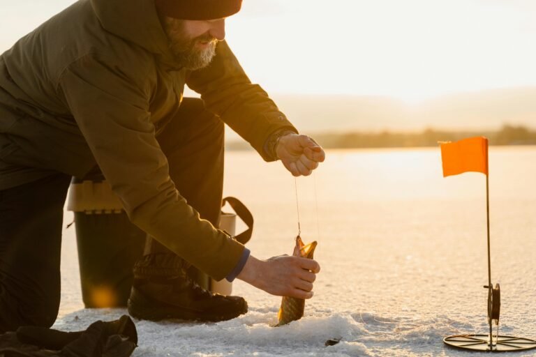 Angler pulling a freshly caught fish from an ice hole during ice fishing in Banff at sunrise, with a tip-up flag and frozen lake in the background.